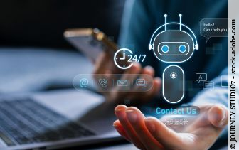 Person am Laptop, die in einer Hand ein Smartphone hält, über der anderen Hand ist die Grafik eines KI Roboters zu sehen